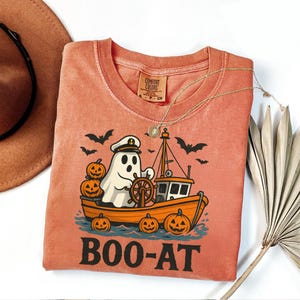 May include: Coral-colored t-shirt with a Halloween-themed graphic. The design features a ghost wearing a captain's hat steering an orange boat, surrounded by pumpkins and bats. The text "BOO-AT" is printed below the boat.