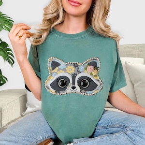 May include: A teal-colored t-shirt featuring a raccoon face design. The raccoon's face is gray and white, with large black eyes and a pearl-beaded outline. Floral accents in yellow, blue, and pink add a decorative touch to the design.
