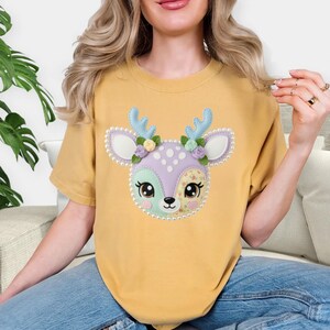 以下が含まれることがあります： マスタードイエローのTシャツ。空想的な鹿の顔のデザインが特徴です。鹿は青い角、花のアクセント、パステルカラーの模様の顔をしています。デザインは真珠のような縁取りで縁取られ、遊び心があり魅力的な美しさを生み出しています。