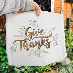 May include: White tote bag featuring the words "Give Thanks" in gold script, with gold and brown botanical illustrations. The bag is held up by a person.