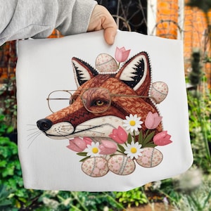 Może przedstawiać: Biała torba z szczegółową ilustracją lisa w okularach, ozdobiona kwiatami i pisankami. Lis ma brązowe futro ze skomplikowanymi wzorami. Różowe tulipany i białe stokrotki otaczają głowę lisa, tworząc fantazyjny design.