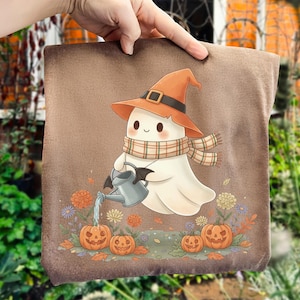 Pode incluir: Uma almofada quadrada castanha com um fantasma de desenho animado usando um chapéu e cachecol laranja, regando abóboras com um regador. O design inclui folhas e flores de outono, ideal para o Halloween.