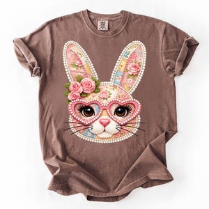 Puede incluir: Camiseta marrón con un diseño de cara de conejo caprichoso. El conejo lleva gafas en forma de corazón y una corona floral. La cara del conejo está adornada con perlas y las orejas tienen un estampado floral de patchwork. Perfecto para Pascua.