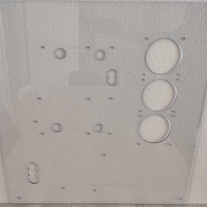May include: A clear, rectangular acrylic panel with various circular cutouts and drilled holes. The panel features three large, evenly spaced circular openings and several smaller holes, suggesting a functional design. The surface has a textured appearance.