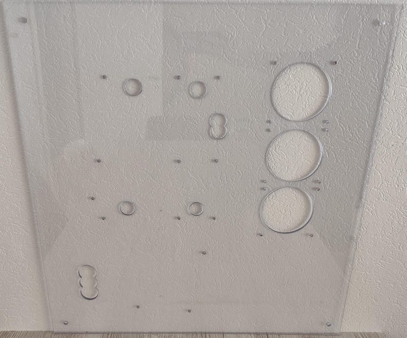 May include: A clear, rectangular acrylic panel with various circular cutouts and drilled holes. The panel features three large, evenly spaced circular openings and several smaller holes, suggesting a functional design. The surface has a textured appearance.