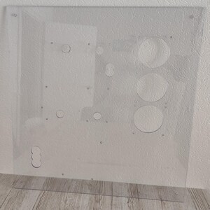 May include: A clear, rectangular acrylic panel with various circular cutouts and small holes. The panel has a series of drilled holes and several larger circular openings. The panel is designed for a specific purpose, likely for electronics or a similar application.