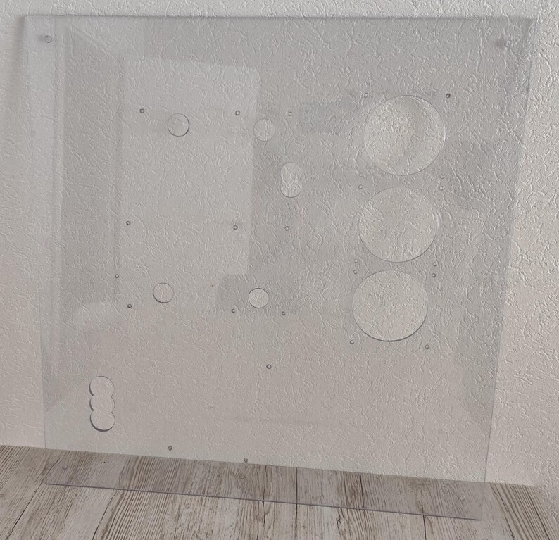 May include: A clear, rectangular acrylic panel with various circular cutouts and small holes. The panel has a series of drilled holes and several larger circular openings. The panel is designed for a specific purpose, likely for electronics or a similar application.