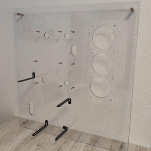 May include: A clear acrylic panel with various circular and rectangular cutouts, along with several small metal pegs and black metal brackets. The panel is mounted with silver hardware, and is designed for display or organization.