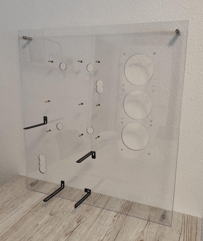 May include: A clear acrylic panel with various circular and rectangular cutouts, along with several small metal pegs and black metal brackets. The panel is mounted with silver hardware, and is designed for display or organization.