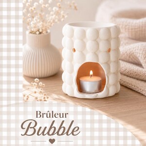 Peut inclure: Un diffuseur d'huiles essentielles en céramique blanche avec un motif de bulles. Une bougie chauffe-plat allumée est à l'intérieur du diffuseur, créant une lumière chaleureuse. Le mot "Bubble" est écrit en marron.