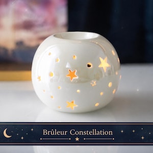 Pode incluir: Um queimador de óleo esférico de cerâmica branca com recortes em forma de estrela. O queimador tem uma abertura circular na parte superior e está rotulado como "Brûleur Constellation" em uma faixa azul marinho com estrelas e uma lua crescente.