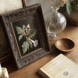 May include: A framed botanical print depicting a plant with spiky seed pods, green leaves, and a pale purple flower. The dark brown frame has decorative carvings. A vintage book and glass vase are also in the image.