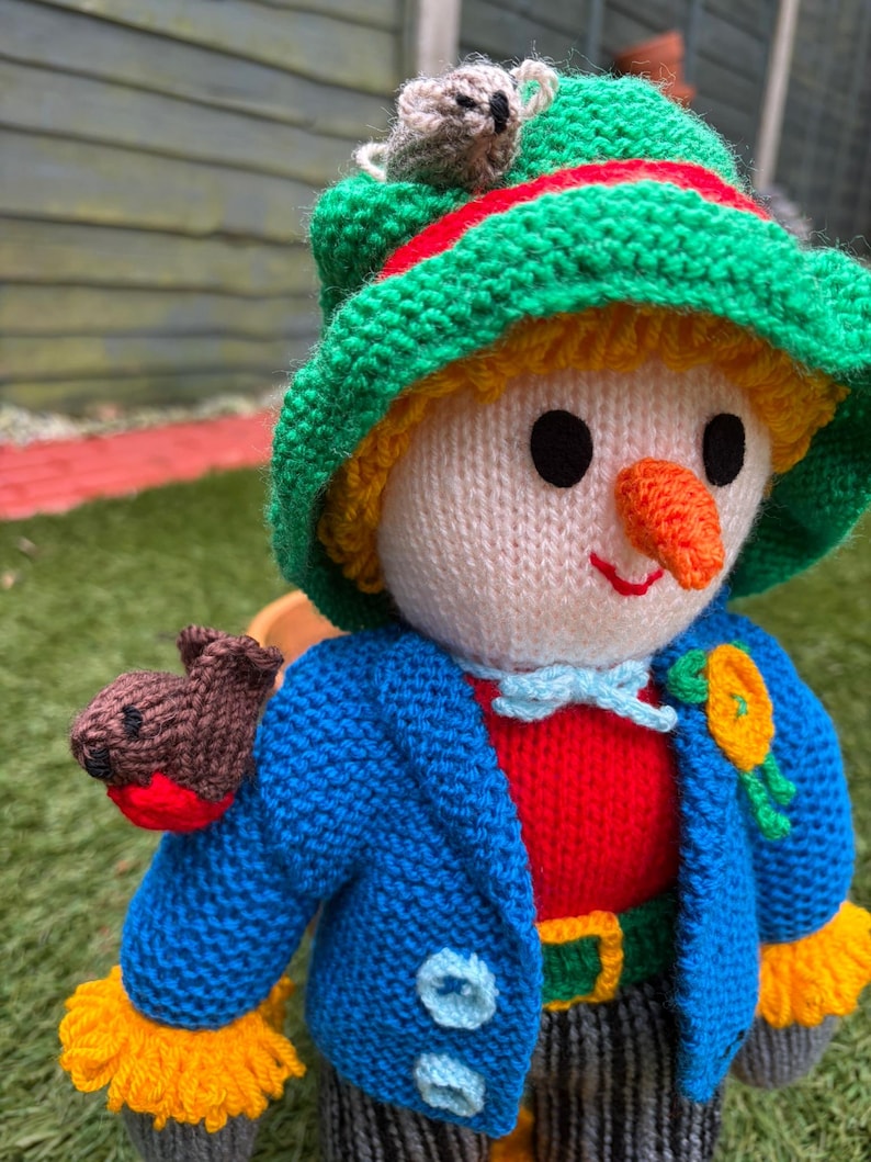 K&ouml;nnte beinhalten: Handgestrickte Puppe mit gr&uuml;nem Hut, rot-gr&uuml;nem Streifen und einem kleinen grauen Koala. Die Puppe tr&auml;gt eine blaue Jacke, ein rotes Hemd und einen gelb-gr&uuml;nen G&uuml;rtel. Ein brauner Vogel sitzt auf der Schulter.