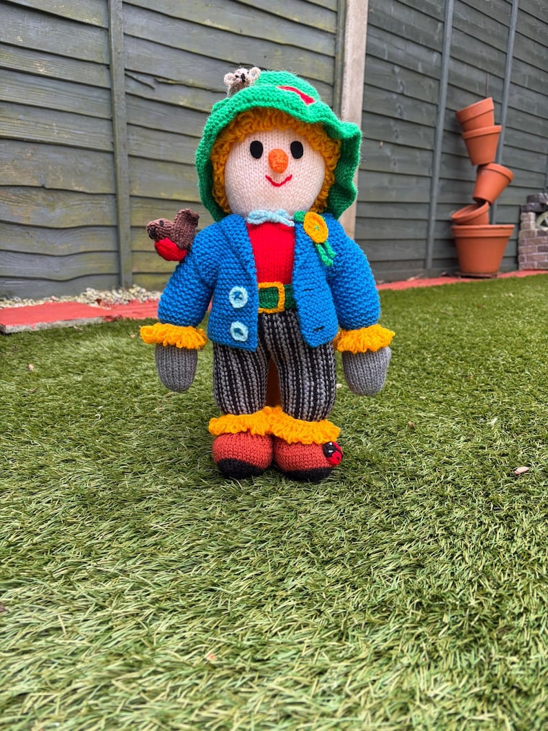K&ouml;nnte beinhalten: Handgestrickte Vogelscheuchenpuppe mit gr&uuml;nem Hut, blauer Jacke und gestreiften Hosen. Die Puppe hat ein l&auml;chelndes Gesicht, ein rotes Hemd und orangefarbene und gelbe Akzente. Ein kleiner Vogel sitzt auf ihrer Schulter.