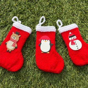 Peut inclure: Trois bas de Noël rouges avec des poignets blancs et des boucles de suspension. Chaque bas présente une application festive différente : un renne, un pingouin et un bonhomme de neige. Les bas sont exposés sur une surface verte.