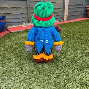 K&ouml;nnte beinhalten: Eine handgestrickte Froschpuppe mit gr&uuml;nem Hut und rotem Band. Die Puppe hat einen blauen Mantel mit zwei hellblauen Kn&ouml;pfen, graue Handschuhe und orangefarbene Verzierungen. Die Beine der Puppe sind gestreift.