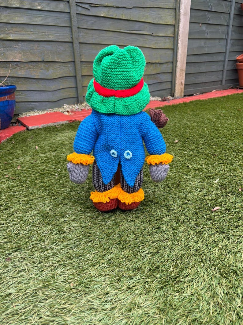 K&ouml;nnte beinhalten: Eine handgestrickte Froschpuppe mit gr&uuml;nem Hut und rotem Band. Die Puppe hat einen blauen Mantel mit zwei hellblauen Kn&ouml;pfen, graue Handschuhe und orangefarbene Verzierungen. Die Beine der Puppe sind gestreift.