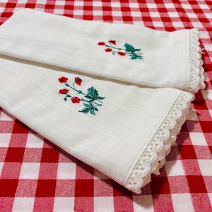 May include: Two white linen hand towels with embroidered red flowers and green leaves. The towels have a decorative lace trim along the edges. The towels are folded and rest on a red and white checkered tablecloth.