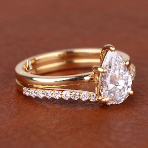May include: A gold ring set consisting of two bands. The primary band features a large pear-cut clear stone held by prongs. The second band is a thin, straight band embedded with a row of small, clear round stones along the visible outer edge.