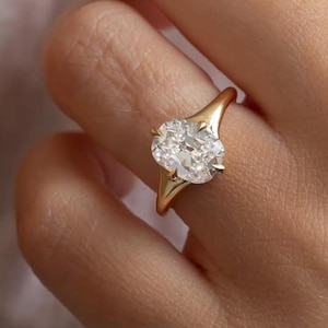 May include: A gold ring featuring a large, oval-cut diamond. The ring's band is a simple, polished gold, and the diamond is set with delicate prongs. The ring is shown on a person's finger, highlighting its elegant design and sparkle.