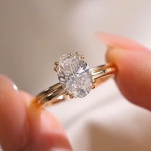 May include: A gold engagement ring featuring a large, oval-cut diamond. The clear diamond is set on a slender gold band. The ring is held up, highlighting its design and sparkle. The ring's design is elegant and classic.