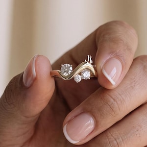Könnte beinhalten: Ein goldener Ring mit einem geschwungenen, wellenförmigen Banddesign, das mit drei runden, klaren Steinen unterschiedlicher Größe besetzt ist. Das Metall hat eine polierte Oberfläche und die Steine sind in einer Gruppe angeordnet.