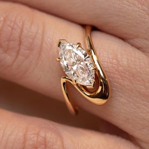 May include: A marquise-cut diamond engagement ring with a unique, swirling gold band. The diamond is set in a four-prong setting, and the band curves around the finger. The ring is a classic and elegant piece of jewelry.