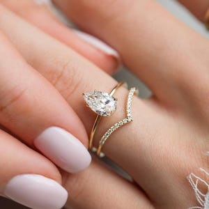 Könnte beinhalten: Ein goldener Verlobungsring mit einem birnenförmigen Diamanten und einem zarten Band. Ein zweiter goldener Ring, V-förmig, ist mit kleinen Diamanten besetzt. Die Ringe werden auf einer Hand mit hellrosa Nagellack gezeigt.