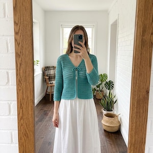 May include: Teal crochet cardigan with a V-neck and three-quarter sleeves. The open-weave design features a tie closure at the bust. The cardigan is paired with a white dress. The image is reflected in a wooden-framed mirror.