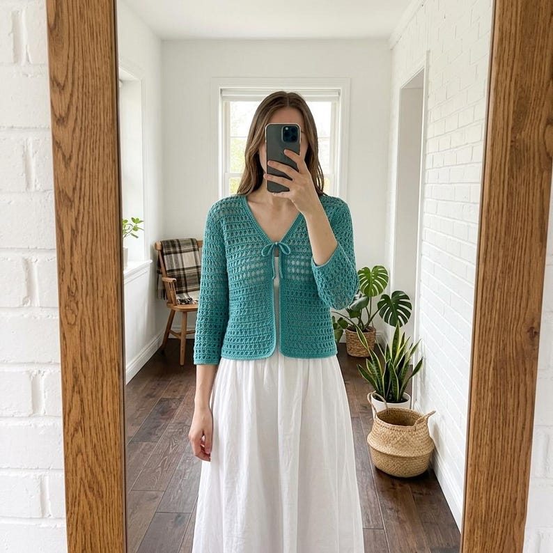May include: Teal crochet cardigan with a V-neck and three-quarter sleeves. The open-weave design features a tie closure at the bust. The cardigan is paired with a white dress. The image is reflected in a wooden-framed mirror.