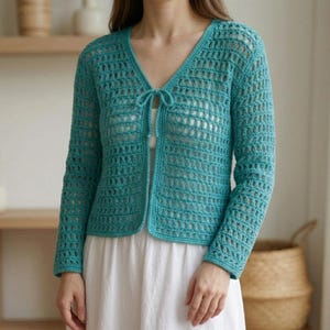 May include: A turquoise, open-weave crochet cardigan with long sleeves and a V-neck. The cardigan has a tie closure at the neckline. The garment is worn over a white dress. The overall style is casual and handmade.