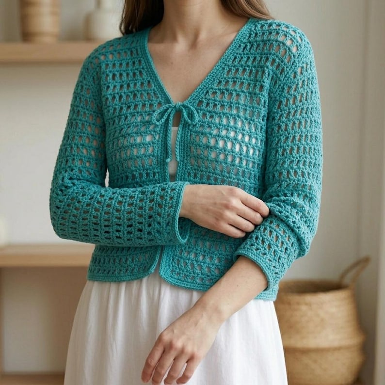 May include: A turquoise crochet cardigan with long sleeves and a V-neckline. The open-weave design features a tie closure at the bust. The cardigan is worn over a white dress. The sleeves are cuffed at the wrists.