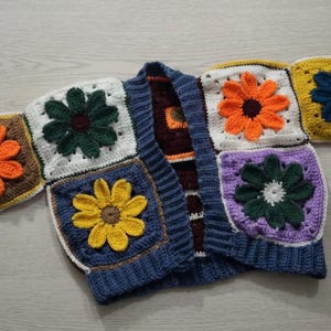 May include: A colorful, handmade crochet cardigan featuring large, square floral motifs. The flowers are in shades of orange, yellow, purple, and green, set against white, brown, and blue backgrounds. The cardigan has a blue ribbed collar and cuffs.