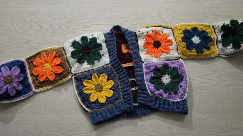 May include: A colorful, handmade crochet cardigan featuring large, square floral motifs. The flowers are in shades of orange, yellow, purple, and green, set against white, brown, and blue backgrounds. The cardigan has a blue ribbed collar and cuffs.