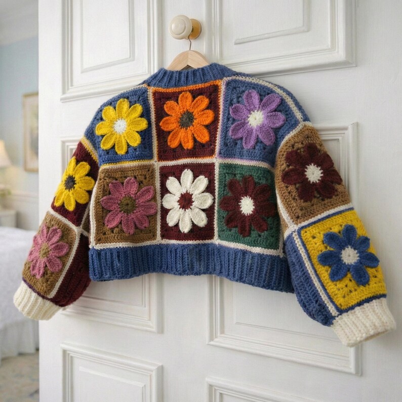 May include: A colorful, crocheted sweater with a patchwork design. The sweater features square panels with various flower designs in yellow, orange, purple, and white. The sleeves and bottom hem are a solid blue, with cream-colored cuffs.
