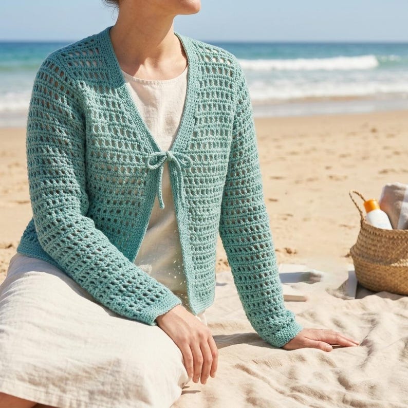 May include: A light turquoise crochet cardigan with long sleeves and a tie closure. The open-weave design allows a glimpse of the cream-colored top underneath. The cardigan is worn on a sandy beach with the ocean in the background.