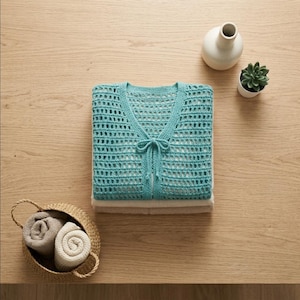 May include: A folded turquoise crochet cardigan with a V-neck and tie closure, resting on a stack of folded fabric. A small woven basket holds rolled towels. A white vase and potted succulent sit nearby on a wooden surface.