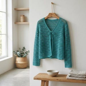 May include: A teal, open-weave crochet cardigan with long sleeves hangs on a wooden hanger. The cardigan has a V-neck and a tie closure at the front. The background includes a wooden bench, a white bowl, and a woven basket.
