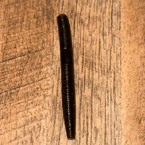Peut inclure: Un leurre de pêche noir et rainuré est posé sur une surface en bois. Le leurre est cylindrique et se rétrécit légèrement vers l'extrémité. Le fond en grain de bois est de couleur brun clair et chaud.