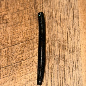 Peut inclure: Un ver de pêche noir avec des paillettes bleues. L'appât en plastique souple est cylindrique avec une texture côtelée. Le ver est présenté sur une surface en bois.