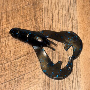 Peut inclure: Un leurre de pêche en plastique souple noir et bleu en forme d'écrevisse. Le leurre a un corps segmenté, deux grandes pinces et est parsemé de points bleus. Le leurre est conçu pour attirer les poissons.