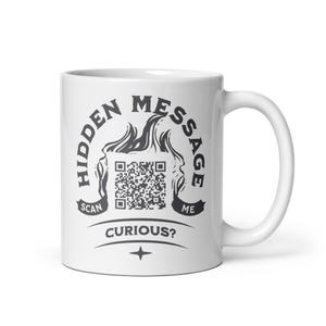 Peut inclure: Un mug en céramique blanche avec un motif graphique gris foncé. L'impression comprend le texte HIDDEN MESSAGE, SCAN ME et CURIOUS? entourant une illustration centrale de code QR encadrée par des flammes, avec une petite étoile à quatre branches.