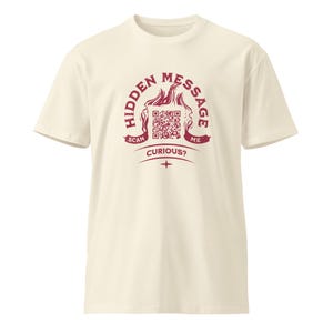 Puede incluir: Camiseta color crema con un gráfico granate. El gráfico presenta las palabras "HIDDEN MESSAGE", un código QR y las frases "SCAN ME" y "CURIOUS?". La camiseta tiene mangas cortas y cuello redondo.