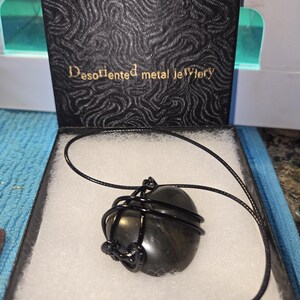 May include: A black pendant necklace with a dark stone wrapped in black wire, presented in a black box. The box has a textured pattern and the words "Desoriented metal Jewelry" in gold lettering.