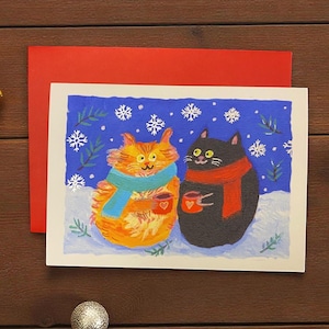 May include: A greeting card featuring two cartoon cats in a winter scene. One orange cat wears a blue scarf, the other black cat wears a red scarf. Both cats hold mugs with hearts, set against a blue snowy background with a red envelope.