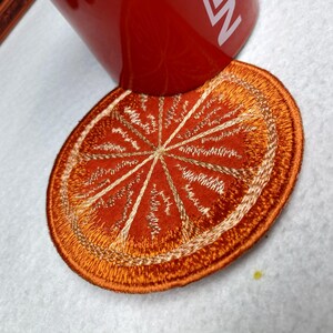 Op de afbeelding: Geborduurde onderzetter in de vorm van een sinaasappelschijf met een gouden rand en interieurdetails. De onderzetter is rond en heeft een levendige oranje kleur, met gouden stiksels die de segmenten van de sinaasappel creëren. Een rode mok met witte tekst rust op de onderzetter.