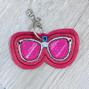 May include: A pink sunglasses keyring with a silver clasp. The sunglasses have pink lenses, a light blue frame, and black outlining. The keyring is made of a stitched material.