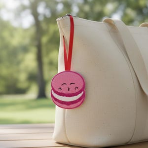 Könnte beinhalten: Ein pinkfarbener, glitzernder Macaron-förmiger Anhänger mit einem lächelnden Gesicht, der an einem roten Band an einer cremefarbenen Tragetasche befestigt ist. Das Macaron hat eine weiße Füllung und ist am Reißverschluss der Tasche befestigt.