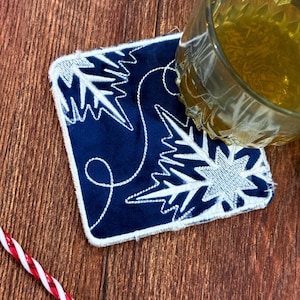 May include: A square, navy blue coaster with white embroidered snowflake designs. A clear glass with a yellow liquid sits on the coaster. The coaster has a white border and is on a wooden surface. A red and white striped straw is in the lower left corner.
