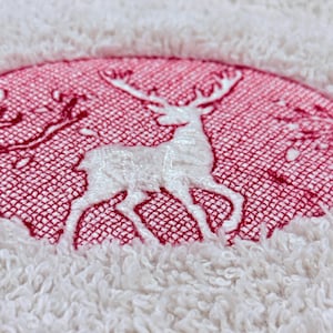 Puede incluir: Primer plano de una toalla blanca con un diseño circular rojo. El diseño presenta un ciervo blanco con grandes astas. La toalla tiene una superficie texturizada y en bucle, y el ciervo está bordado con finos detalles.